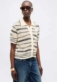 Γυναίκα που φοράει ριγέ πλεκτό μπλουζάκι με κουμπιά και κοντά μανίκια, μπλε τζιν, μοβ γυαλιά ηλίου, χρυσούς κρίκους σκουλαρίκια και χρυσό βραχιόλι.