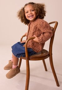 Cardigan en laine marron, jupe en jean, chaussettes roses et bottes beige portés par une fille assise sur une chaise en bois sur un fond neutre.