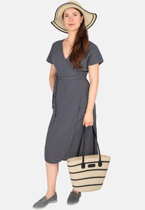 Frau trägt graues Wickelkleid, breitkrempigen Hut, graue Loafers und hält eine beige-schwarz gestreifte Tragetasche auf weißem Hintergrund.