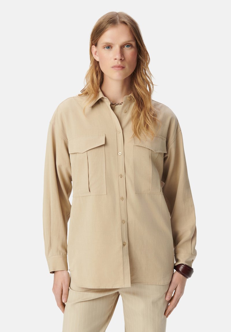 Beige langwerpig shirt met twee borstzakken, knoopsluiting aan de voorkant en een relaxte pasvorm. Glad textuur, kraagontwerp en subtiele glans.