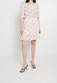 Abito bianco con motivo floreale rosso, scollo a V, maniche corte a sbuffo, vita arricciata e orlo arricciato. Modella che indossa sandali verde con tacco.