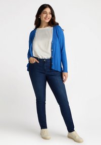 Cárdigan azul con botones dorados sobre una blusa blanca, combinado con jeans oscuros y zapatillas ligeras. Textura suave, corte casual y diseño en capas.