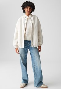 Witte fleecejas met drukknoopjes, gedragen over een witte geribde trui. Lichtblauwe wijde jeans en beige sneakers maken de outfit compleet.