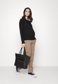 Top preto de manga longa com ribanas, calças beige, bolsa tote preta com bolso com fecho, combinado com ténis pretos. Modelo está de pé contra um fundo branco.