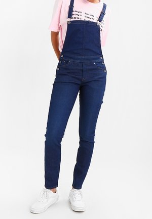 Personne portant une salopette en denim bleu foncé avec un t-shirt rose et des baskets blanches, debout devant un fond blanc uni.