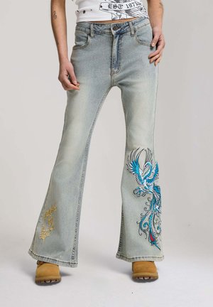Light blue flared jeans with colorful phoenix embroidery on right leg and gold script on left leg, worn with tan boots.