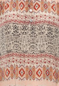 Haut en tissu à motifs avec des dessins floraux et géométriques ornés. Couleurs incluant beige, noir, rouge et orange. Détails avec des boutons présents.