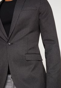 Blazer rayé sombre en tissu texturé, comportant une fermeture à un bouton, des revers crantés et une poche poitrine.