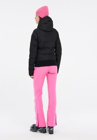 Schwarze Pufferjacke, cropped Länge, strukturierter Stoff. Leuchtend pinke, dehnbare Skihosen mit Reißverschlüssen an den Knöcheln. Gestrickte pinke Mütze.