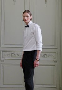 Cămașă albă cu mânecă lungă și cravată neagră, asociată cu pantaloni negri la comandă, pe un fundal cu un perete vintage cu detalii ornamentale.