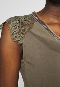 Olive green garment featuring lace trim on the shoulder, mesh overlay at the neckline, and a smooth fabric finish with a fitted shape.