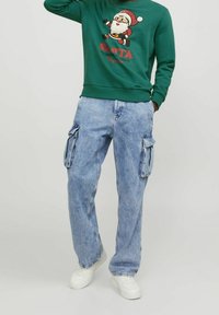 Groene sweatshirt met een Santa-afbeelding en rode tekst, gecombineerd met lichtblauwe cargobroeken en witte sneakers. Casual fit en relaxed ontwerp.