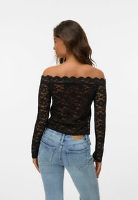 Zwarte kanten off-the-shoulder top met lange mouwen, gegolfde halslijn en bloemenpatroon, gecombineerd met lichtblauwe jeans.