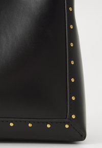 Sac à main noir en cuir lisse, ornée d'ét studs dorés sur le bord, avec une forme structurée et des coins angulaires.