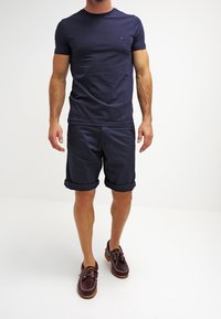 T-shirt blu navy a maniche corte, pantaloni corti blu navy arrotolati e scarpe da barca marroni. Il tessuto sembra liscio, con un logo discreto sulla maglietta.