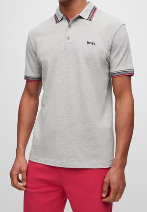 Homme portant un polo gris clair avec des bordures roses et noires, logo noir « BOSS » sur la poitrine, et pantalon rose vif, debout devant un fond blanc.