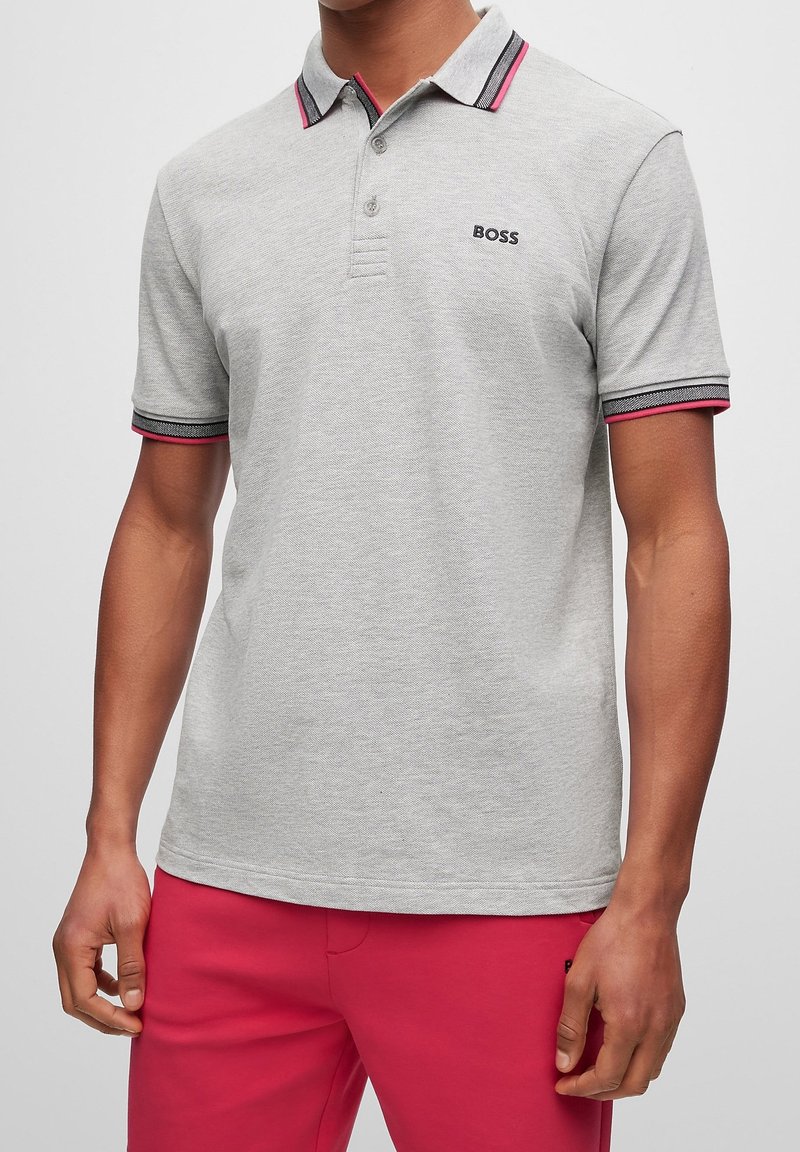 Homme portant un polo gris clair avec des bordures roses et noires, logo noir « BOSS » sur la poitrine, et pantalon rose vif, debout devant un fond blanc.