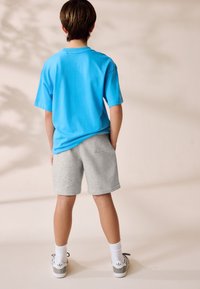 Tricou albastru cu mânecă scurtă, pantaloni scurți gri, șosete albe și adidași cu accente gri. Țesătura pare moale, cu o croială lejeră.