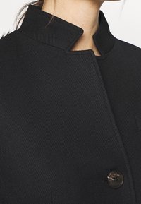 Black coat with a high collar, textured fabric, and a single button closure featuring a logo accent. Close-up view highlights design details.