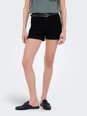 Femeie care poartă pantaloni scurți negri cu tiv răsucit și curea neagră, top verde și pantofi slip-on negri cu detalii decorative.