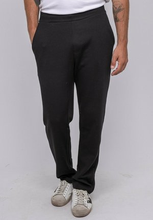 Homme portant un pantalon de survêtement noir, des baskets blanches et un t-shirt blanc, debout les mains dans les poches devant un fond uni.