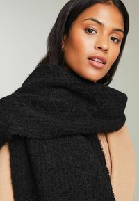 Sort fleece-scarf med en struktureret overflade, draperet om halsen, der har et blødt udseende og betydelig bredde.