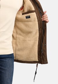 Ruskea samettitakkki, jossa on beigeä fleecettä vuorina. Takissa vetoketju ja nepparit. Sisäpuolella labeli, helmassa säädettävä kiristysnauha.