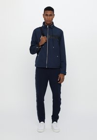 Mørkeblå zip-up jakke med glidelås foran, justerbar hette og svart stripe detalj; kombinert med matchende mørkeblå joggebukser og hvite sneakers.