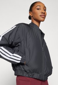 Črna, lahka bomber jakna z zadrgo spredaj, elastičnimi manšeti in belimi trakom s tremi črtami na rokavih. Gladka, sijoča tekstura.