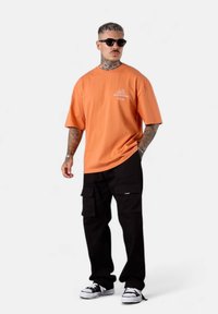 T-shirt ample en coton orange avec un petit logo blanc, associé à un pantalon cargo noir avec des poches et des baskets noir et blanc.