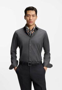 Mand iført en mørkegrå skjorte med knapper, sorte bukser, ternet halstørklæde og sort bælte, stående med hænderne i lommerne mod en hvid baggrund.