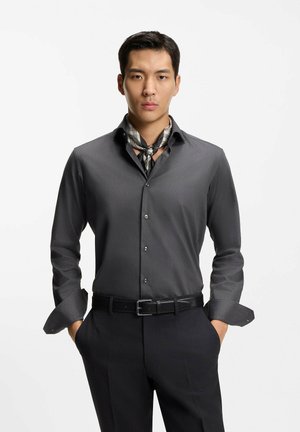 Homme portant une chemise gris foncé à boutons, un pantalon noir, une écharpe à carreaux autour du cou et une ceinture noire, se tenant les mains dans les poches devant un fond blanc.