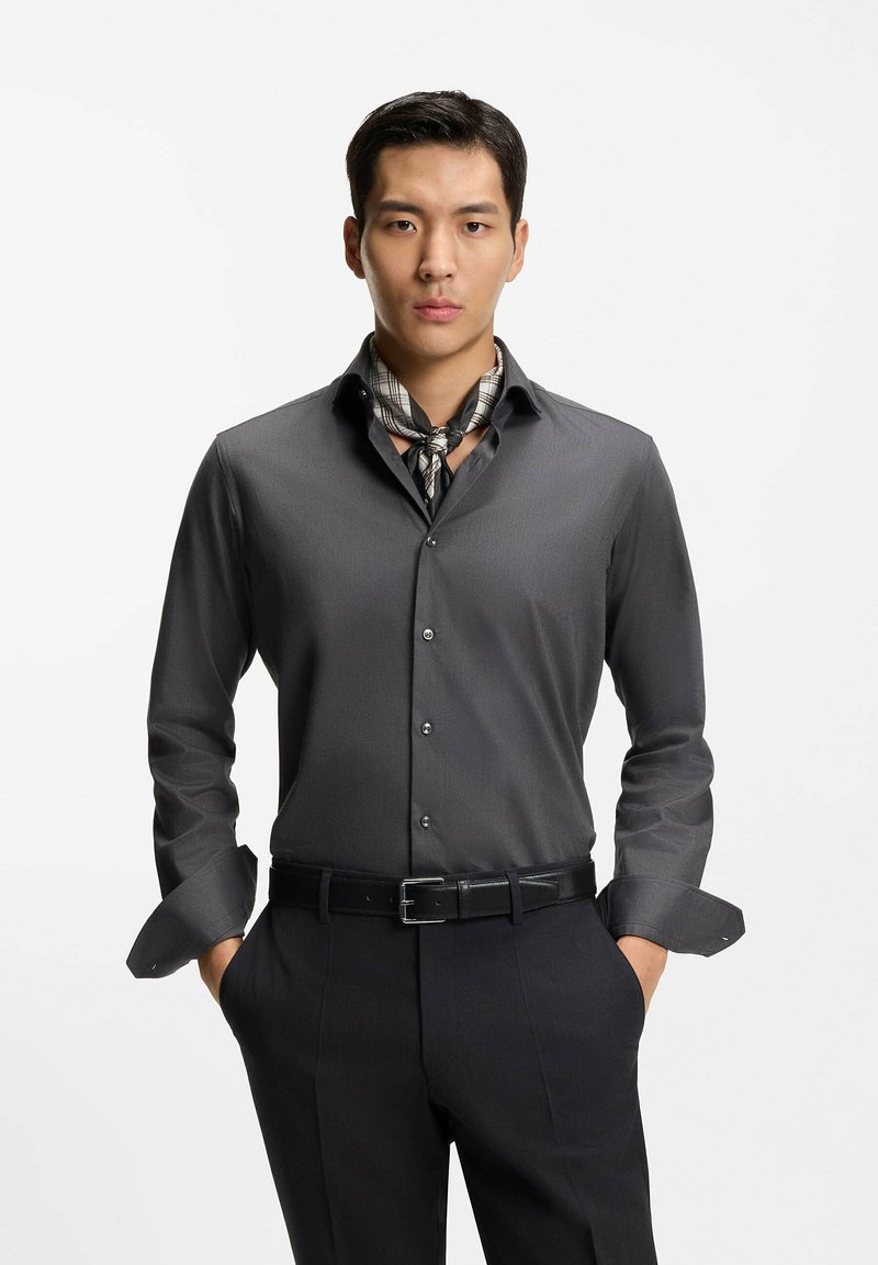 Mand iført en mørkegrå skjorte med knapper, sorte bukser, ternet halstørklæde og sort bælte, stående med hænderne i lommerne mod en hvid baggrund.