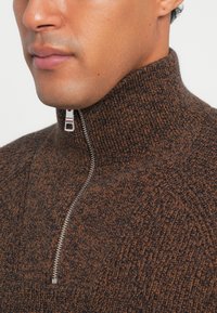 Brun struktureret striktrøje med høj krave og sølv lynlåsdetalje. Har et subtilt mønster og en blød overflade.