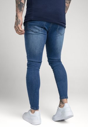 Homme tatoué portant un jean skinny bleu foncé et des baskets blanches, debout les jambes légèrement écartées devant un fond uni.