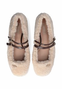 Een paar beige donzige ballerina's met bruine dubbele banden en kleine gouden decoratieve logo's op een witte achtergrond.