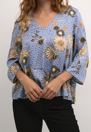 Femme portant un chemisier ample bleu clair à motifs floraux beige et noirs, assorti d'un pantalon noir, debout les mains jointes.