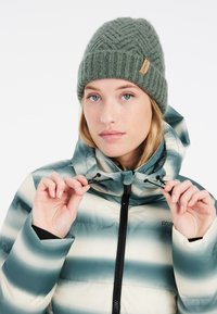 Grüne Strickmütze mit strukturiertem Muster und einem Etikett. Gepolsterte Jacke mit horizontalen Streifen in Türkis und Creme, mit hohem Kragen und Kordeln.