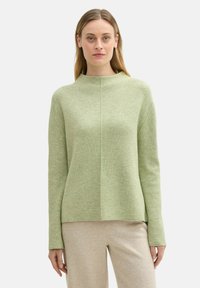 Hellgrüner Pullover mit hohem Kragen, gerippter Textur und flachen Nähten. Lange Ärmel und eine lockere Passform, kombiniert mit hellbeigen Hosen.