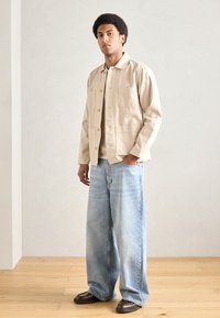 Beżowa bawełniana kurtka z zapięciem na guziki i dwoma kieszeniami na piersiach; w połączeniu z jasnymi, szerokimi dżinsami i brązowymi butami. Neutralne tło.