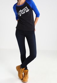 Svart och blå långärmad skjorta med 'Vans' logotyp, i kombination med mörka jeans och ljusbruna snörstövlar. Har färgkontrast på ärmarna och textil med struktur.