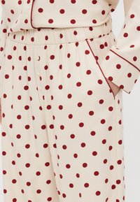 Polyester pyjamasæt med en beige baggrund og burgunder prikker, elastisk talje og kontrastpiping langs manchetterne og kraven.