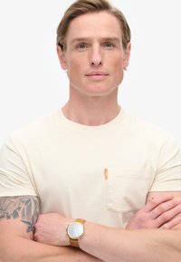 Creme kortærmet t-shirt med en enkelt brystlomme, der har et orange mærke. Modellen bærer et guldur med et brunt læderrem.