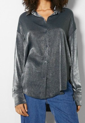 Frau trägt ein langärmliges, glänzendes graues Hemd mit Knöpfen und blaue Jeans, steht vor einem schlichten hellen Hintergrund.