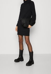 Robe pull noire en maille avec des détails texturés sur les épaules, associée à une pochette noire et des bottines noires épaisses. Tissu lisse, coupe ajustée.