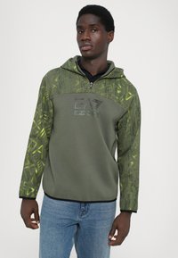 Sweatshirt verde-azeitona com fecho de correr, design texturizado e padrões geométricos amarelos nas mangas. Logótipo preto na parte do peito.