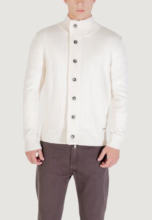 Hombre vistiendo un cárdigan de punto de color crema con cuello alto y pantalones marrón oscuro, de pie frente a un fondo liso.