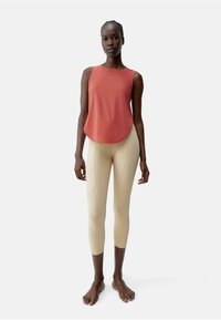 Rode mouwloze top met afgeronde zoom, gecombineerd met strakke beige leggings met naaddetails en een gladde textuur.