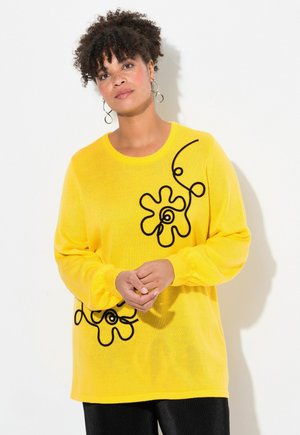 Pull en maille jaune avec un motif floral noir, manches longues et col rond. Texture lisse et coupe ample, accentuée par des détails ludiques.