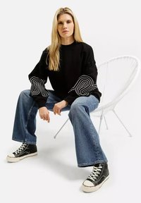 Blondynka siedząca na białym krzesle z siatki, ubrana w czarną bluzę z białymi wzorami, niebieskie dżinsy i czarne sneakersy wysokie.